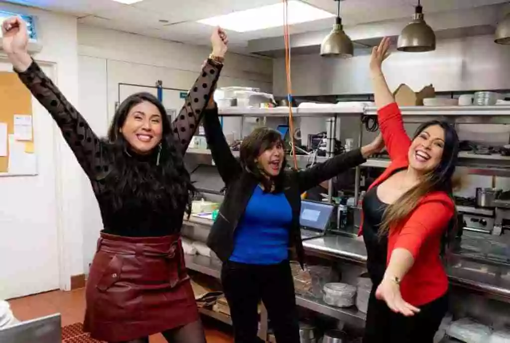 Three women celebrating in a kitchen, raising their arms in joy